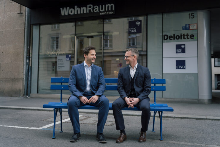 Zwei WohnRaum-Berater der Hypo Tirol auf der hypoblauen Bank vor dem WohnRaum-Kompetenzcenter