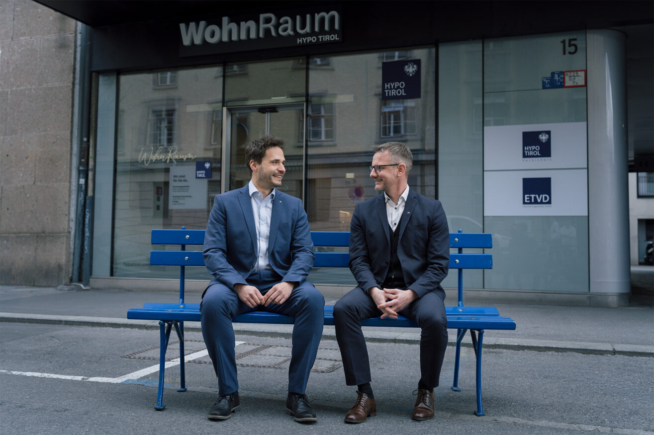 Zwei WohnRaum-Berater der Hypo Tirol auf der hypoblauen Bank vor dem WohnRaum-Kompetenzcenter