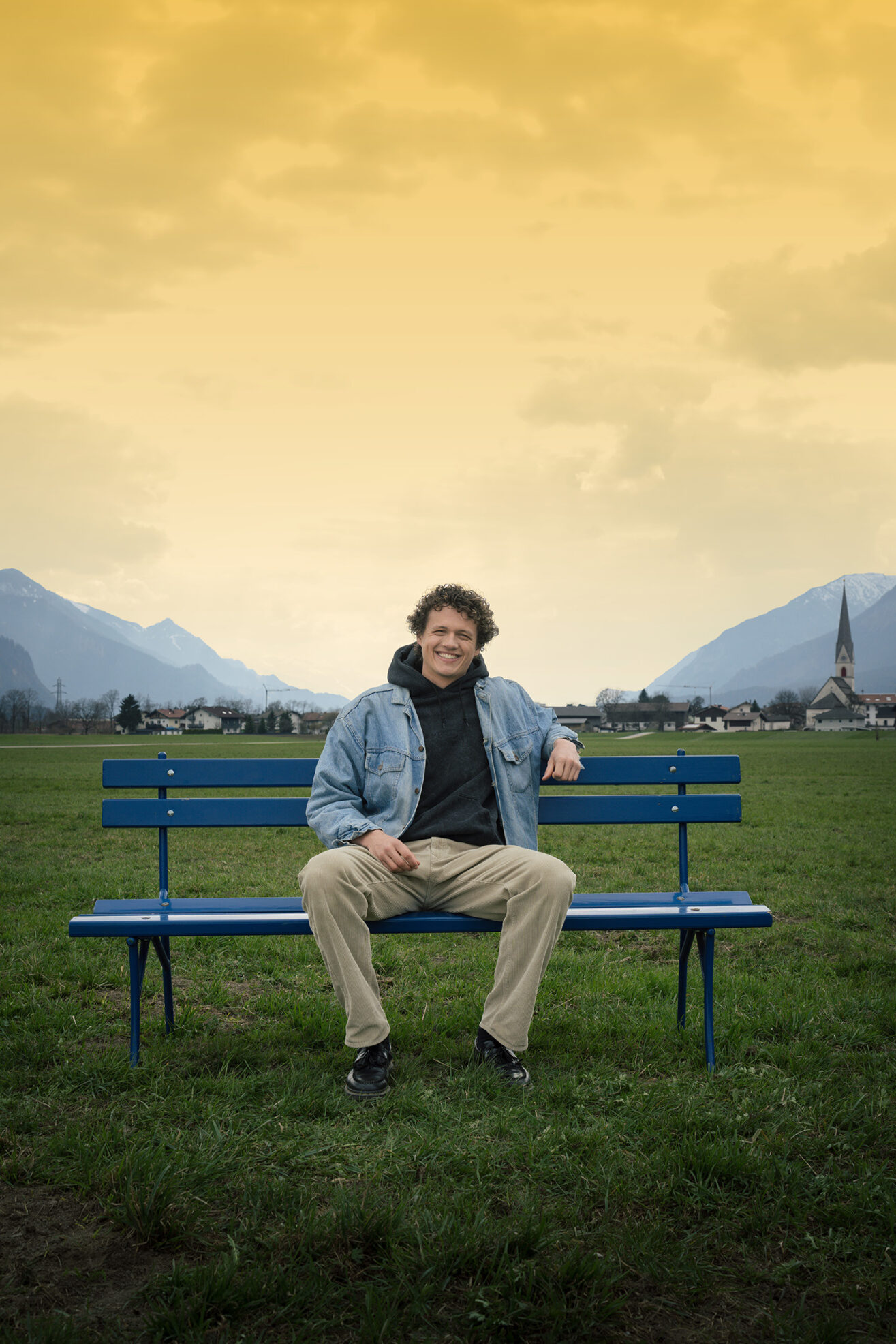 Junger Mann sitzt lächelnd auf der hypoblauen Parkbank auf einer grünen Wiese, der Himmel ist gelb eingefärbt.