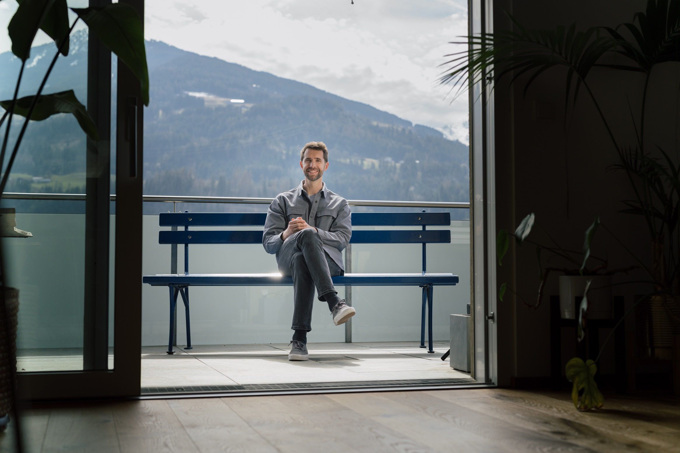 Mann sitzt auf der hypoblauen Parkbank, die auf einer Dachterrasse steht. Im Hintergrund ein Berg.