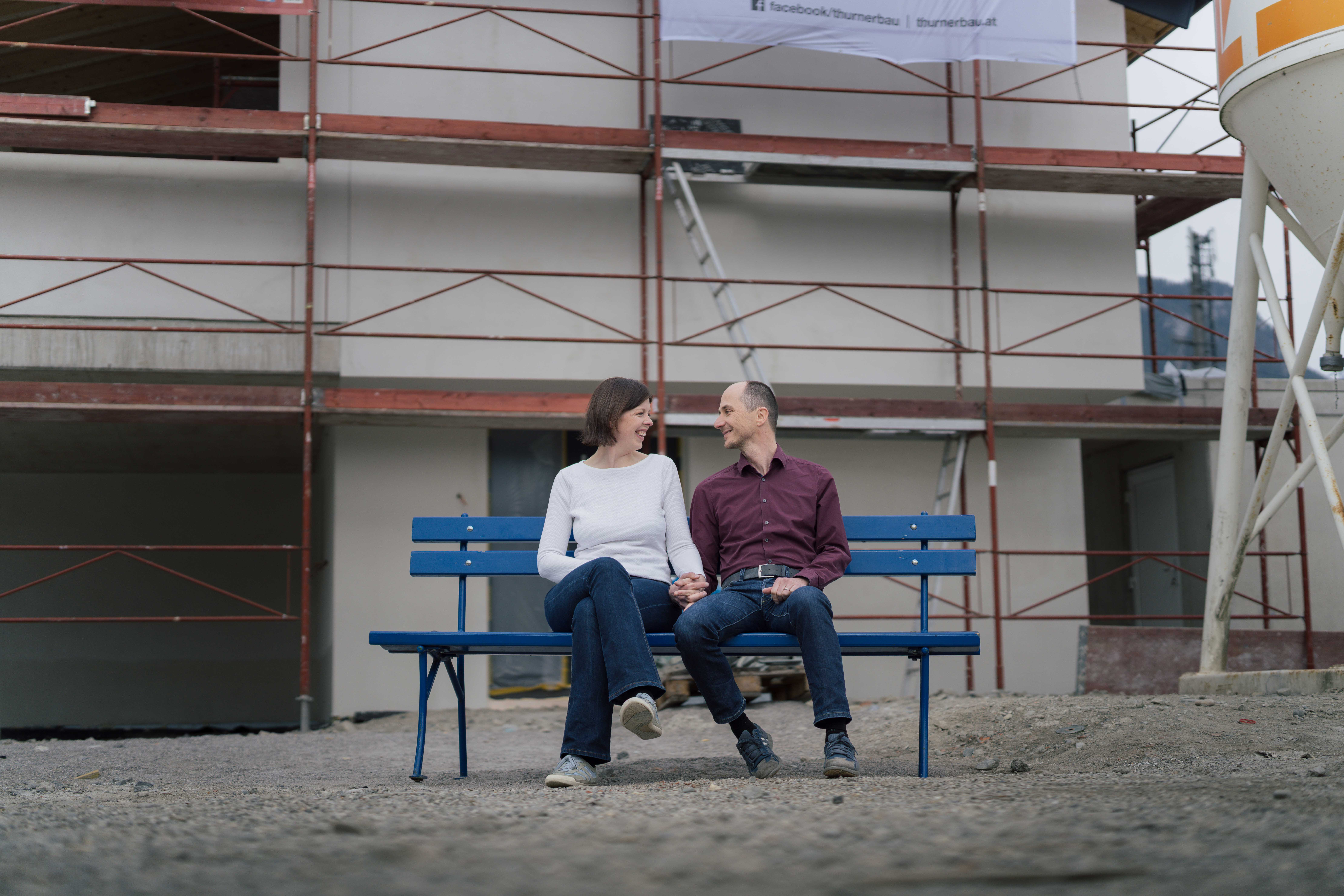 Ein Paar sitzt händchenhaltend auf der hypoblauen Parkbank, die vor einem Haus mit Gerüst steht.